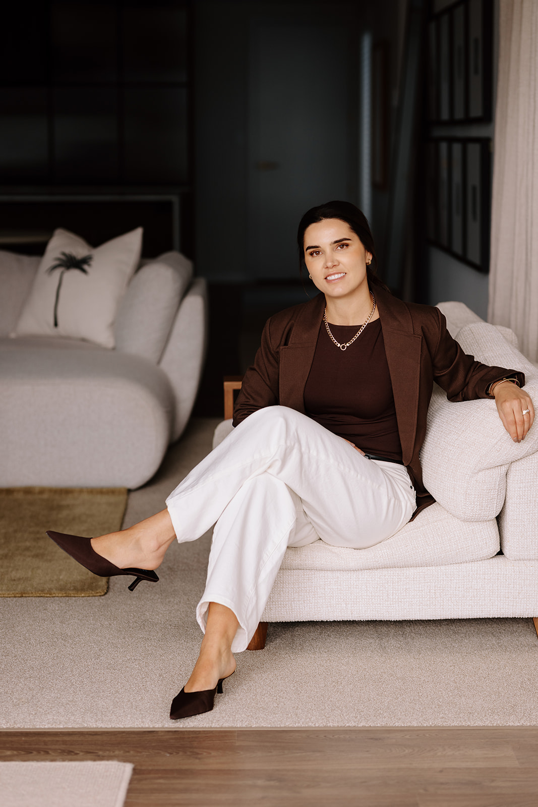 Professional woman in brown blazer sitting on cream sofa in modern bedroom